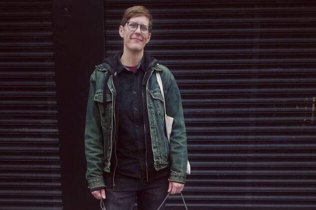 Rian in Dublin, after a massage. A smiling person with short strawberry blond hair and vintage denim jacket stands in front of a black background on a city street.